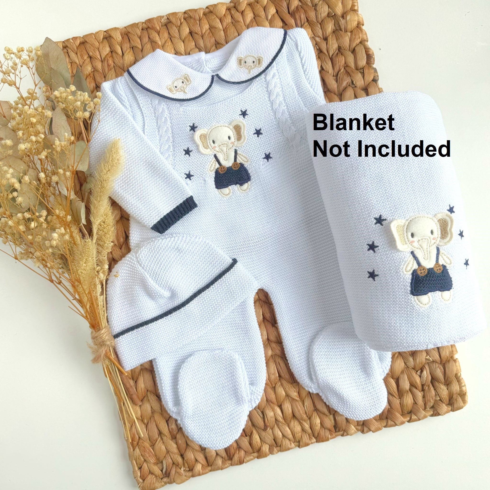 A three-piece knitted baby outfit featuring white with navy details and a cute little elephant design, laid out on a wicker surface.