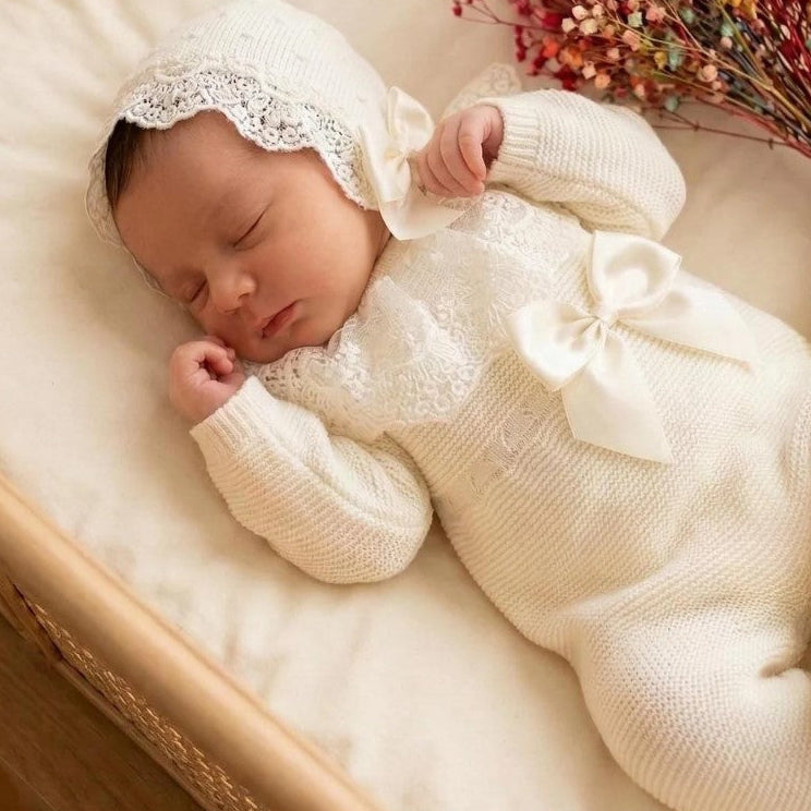A sleeping newborn baby wearing a cream-colored ribbed footed romper with a lace-trimmed bonnet and a large bow on the chest, lying on a cream cushion inside a woven rattan bassinet with dried flowers nearby.