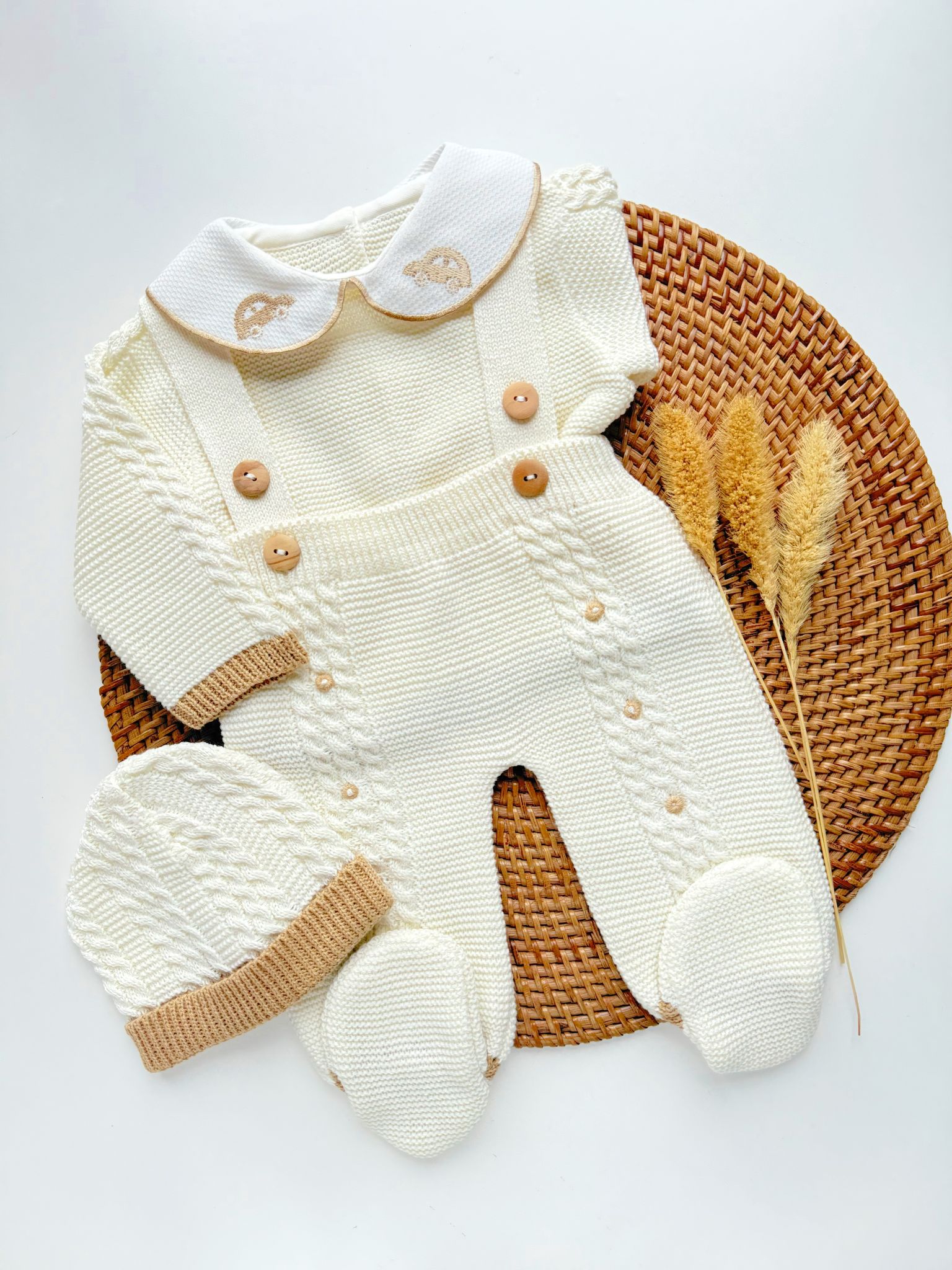 Knitted baby outfit with white sweater and beige overalls on a woven mat.