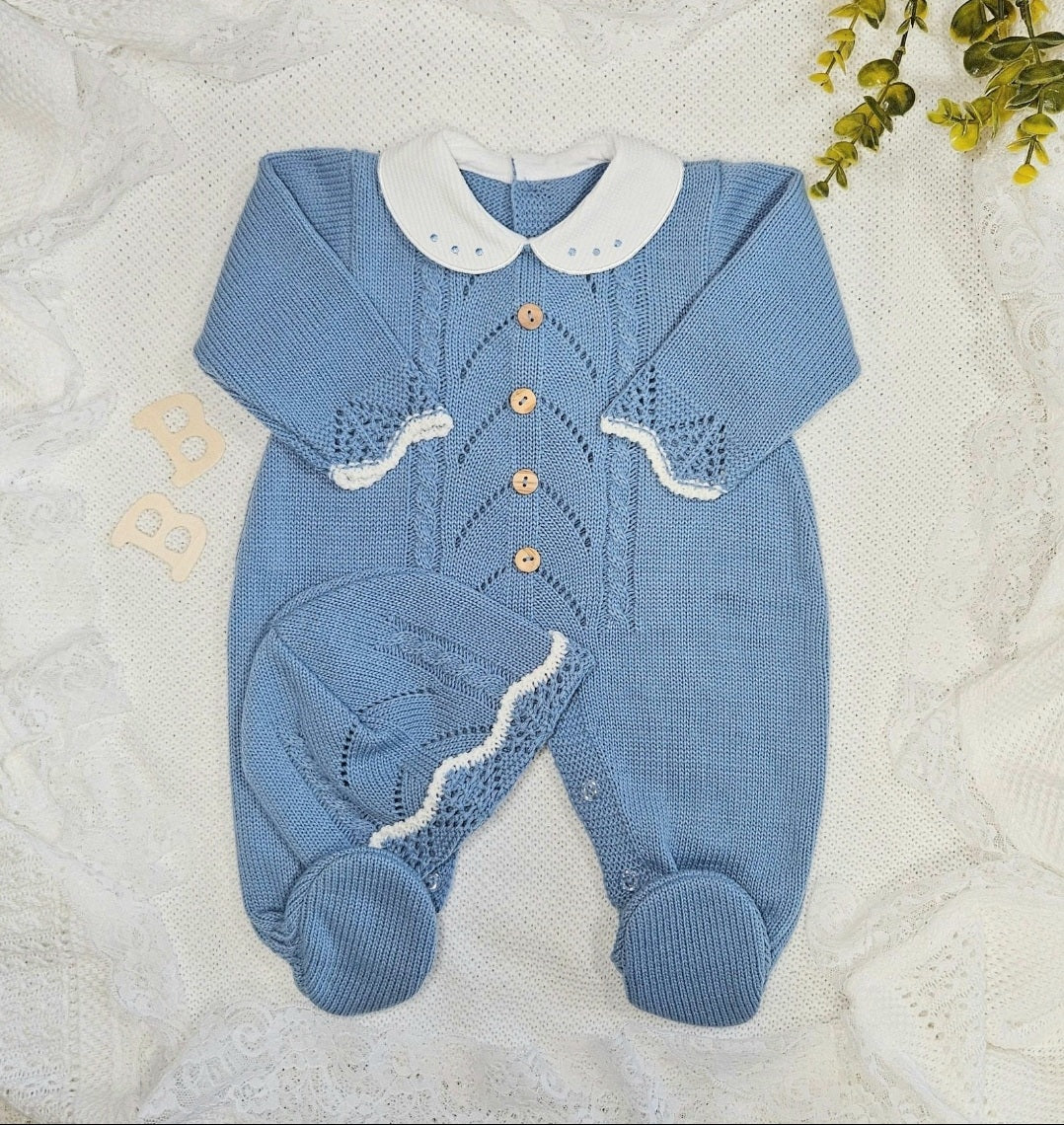 A knitted blue baby outfit with a white collar and blue buttons, accompanied by a matching bonnet, laid out on a white textured background.
