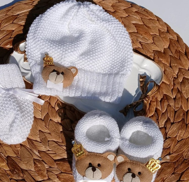 A baby gift set including a white knit bonnet with a teddy bear face and gold crown, matching booties, and a white mitt, arranged on a woven straw mat.