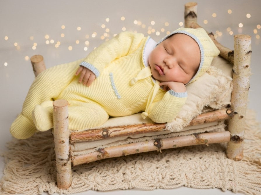 Newborn baby in a yellow outfit sleeping on a small wooden bed with blurred lights in the background