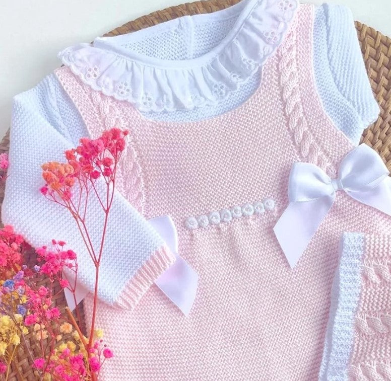 Pink knitted dress with white ruffled collar and bow, surrounded by pink flowers.