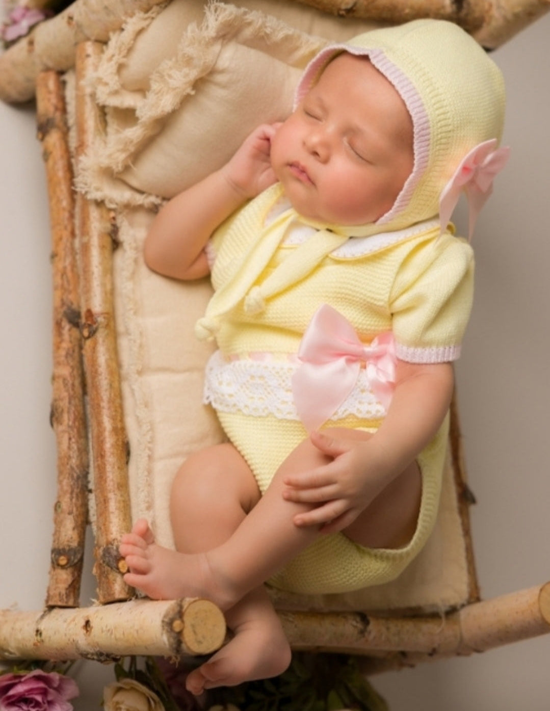 Newborn baby in a yellow outfit and bonnet lying on a wooden crib with flowers around.