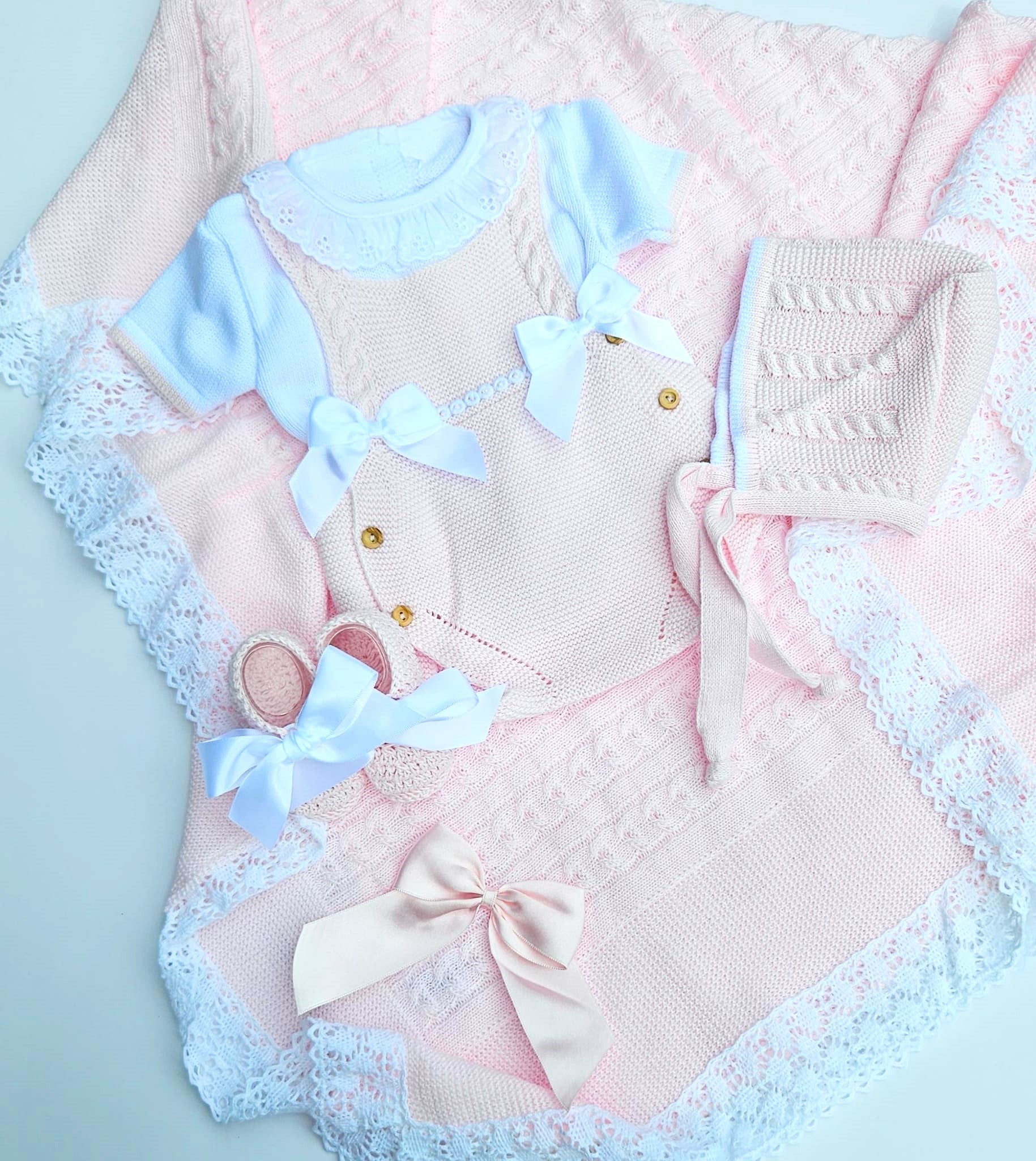 A baby girl's knit outfit set laid flat on a pale blue background, including a light pink and blue short-sleeve romper with lace trim, a matching pink knitted hat, a pink knitted boot, and a pink knitted bonnet with a large bow.