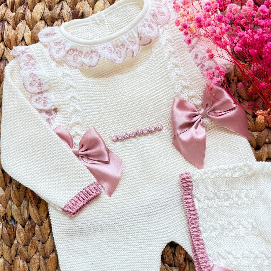White baby outfit with pink bows on a woven mat with pink flowers.