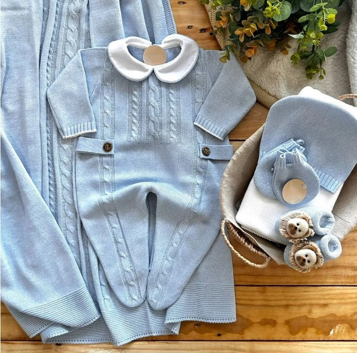A light blue baby layette set including a romper, hat, mittens, and a blanket laid out on a wooden surface.