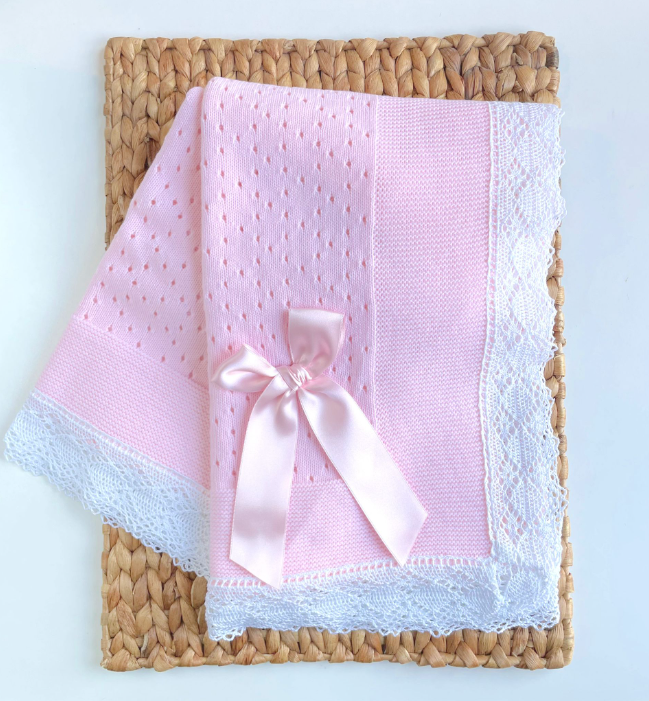 A folded pink and white knitted baby blanket with a white lace edge, presented on a wicker surface. soft baby girl knit blanket