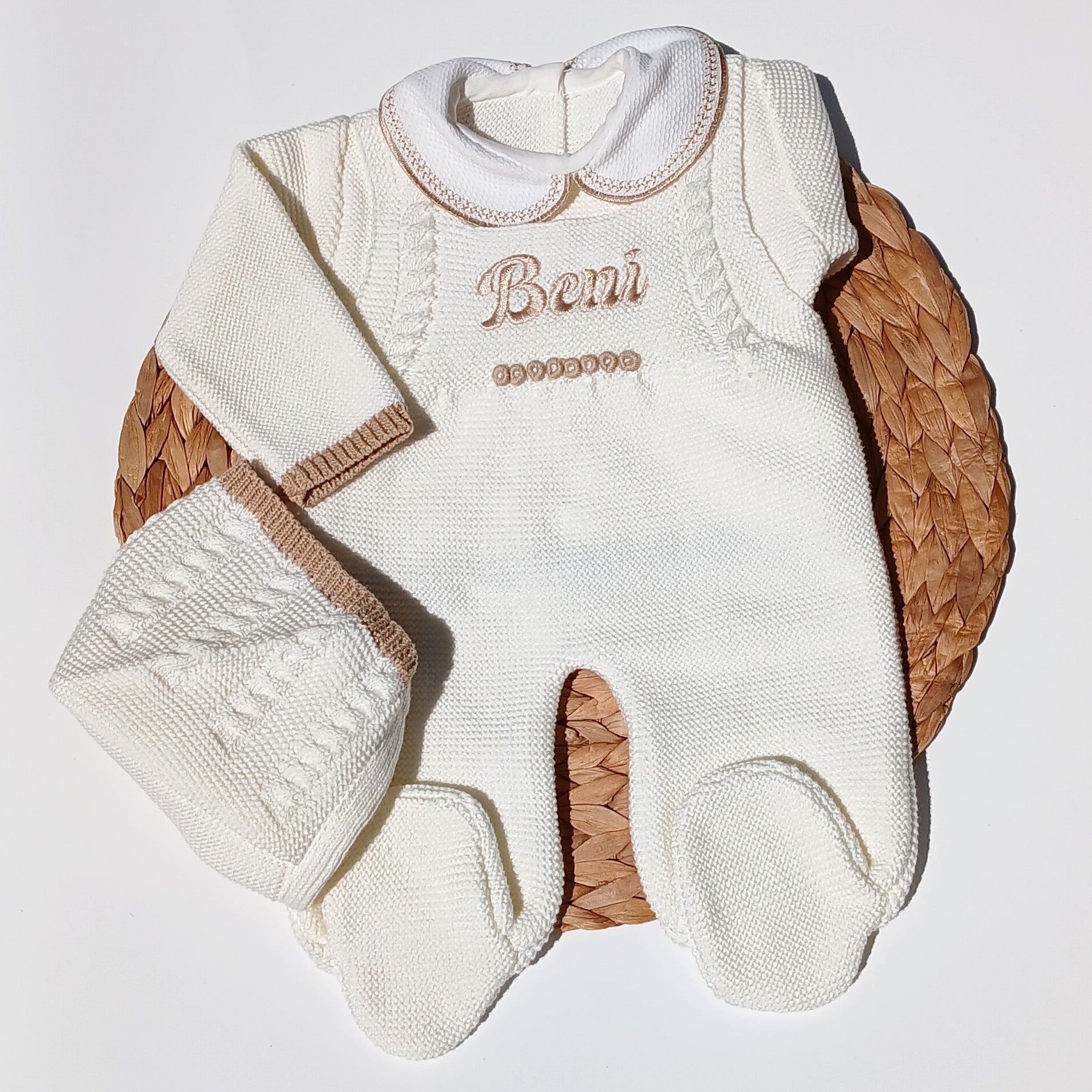 An off-white knit baby footed one-piece romper with 'Beni' embroidered on the chest, displayed on a woven brown mat against a white background.