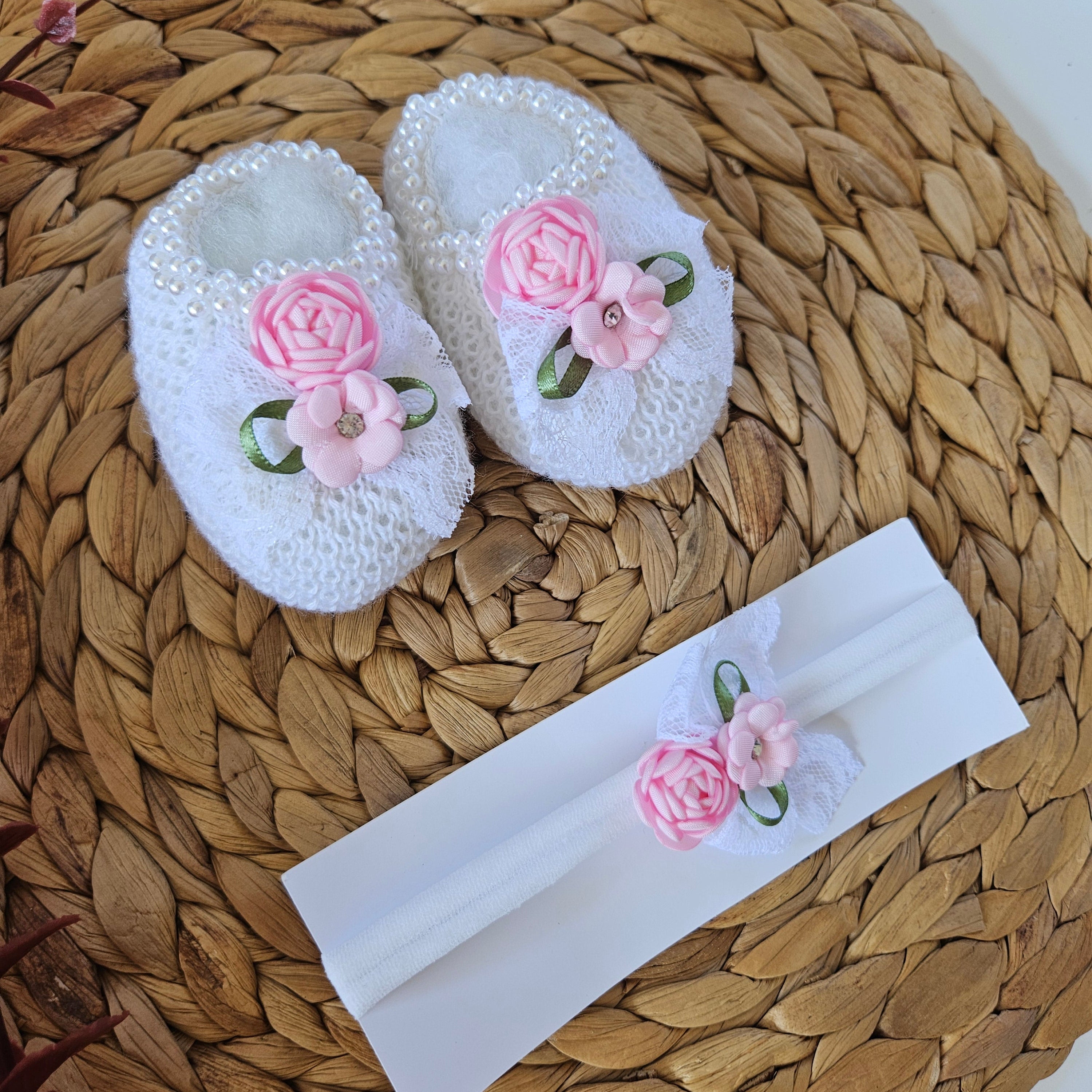 Handmade knitted baby bootie in white with pink flower details alongside a matching headband, placed on a woven mat.