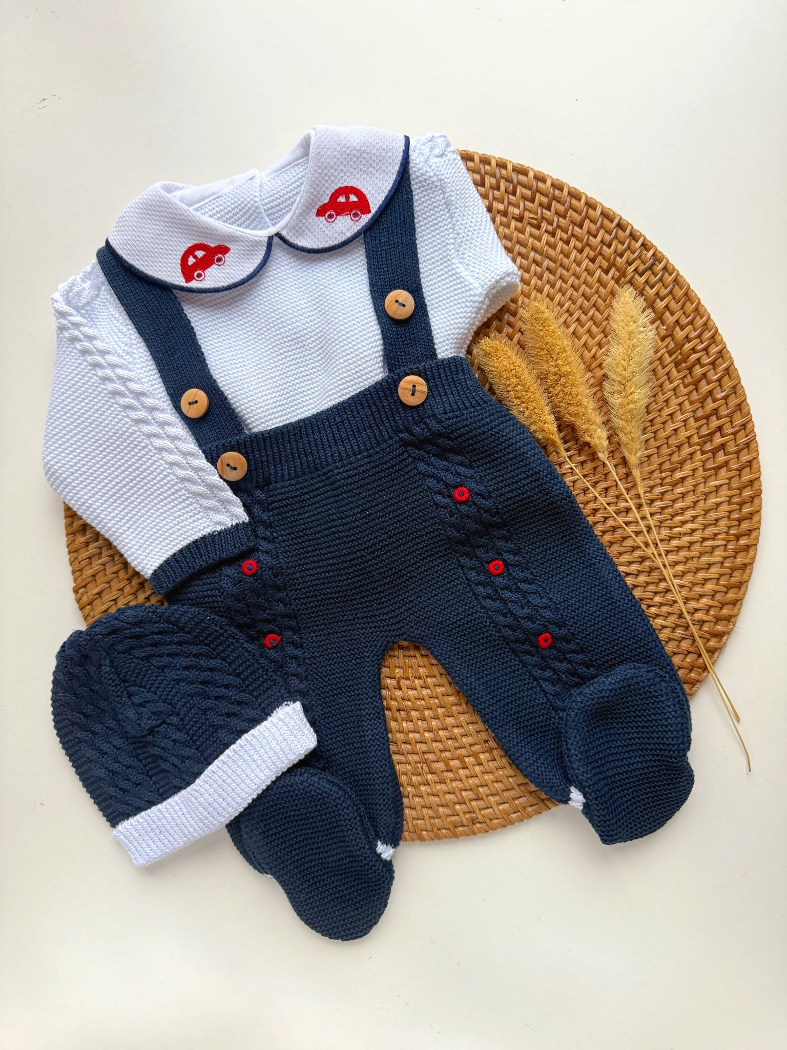 Baby outfit with navy blue knitted overalls, white shirt with red car design, and matching hat on a woven mat.