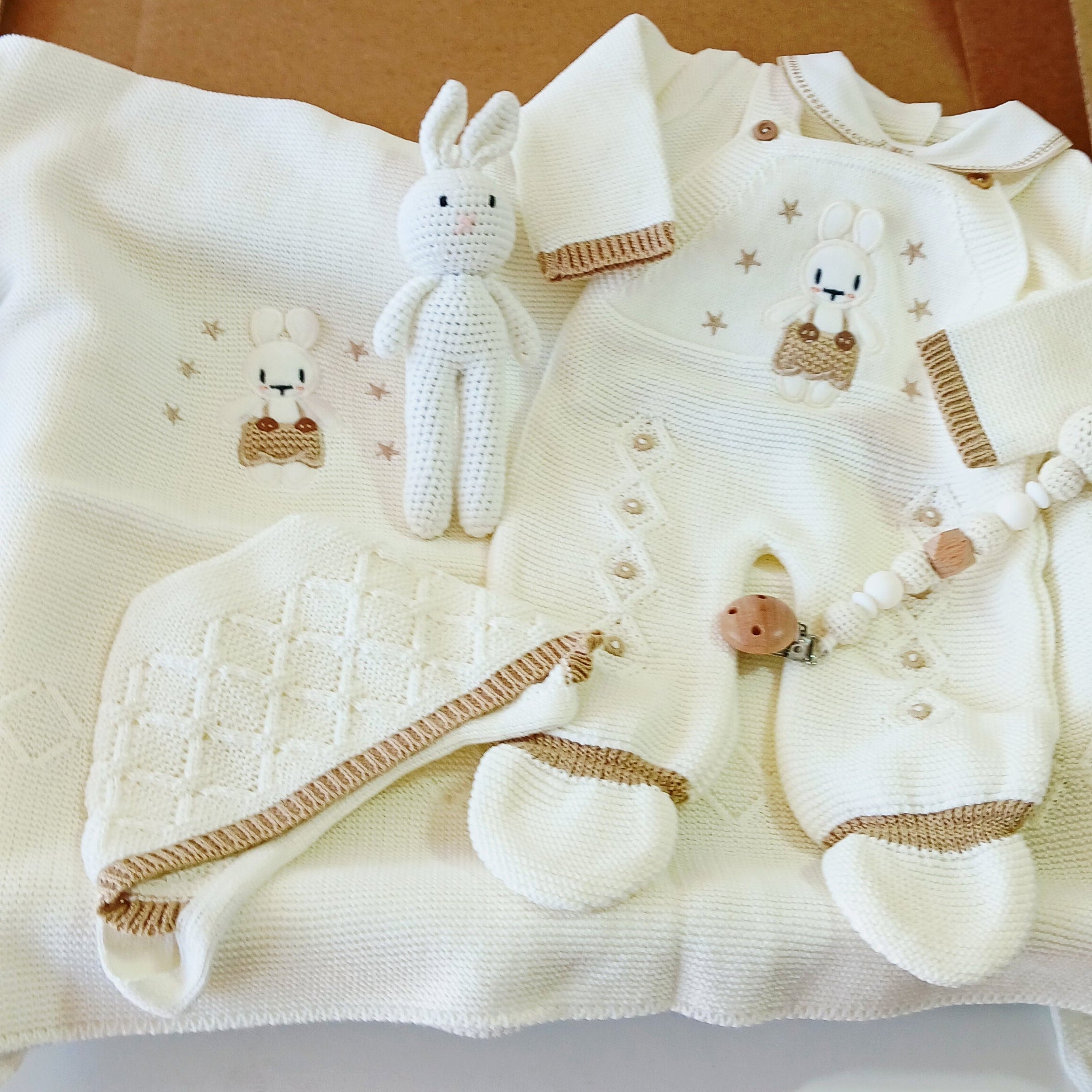 Collection of baby clothes and accessories on a white blanket with a cardboard box in the background.
