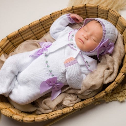 Newborn baby in a white outfit with purple accents lying in a woven basket. Pink baby romper with floral details, newborn girl knit going home outfit with flowers embroidered details. the baby outfit is in pink. Baby girl knit coming home outfit pink and sweater in white, collar with handmade flowers