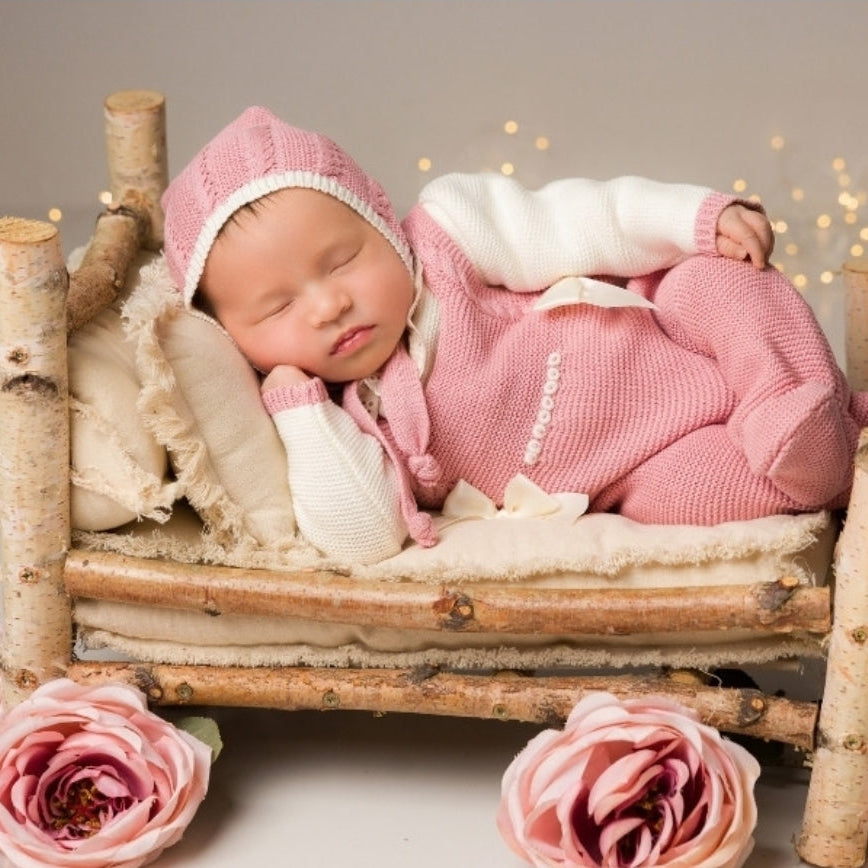 Newborn baby in pink outfit lying on a wooden bed with pink flowers around , newborn knit going home outfit, newborn baby girl coming home knit outfit