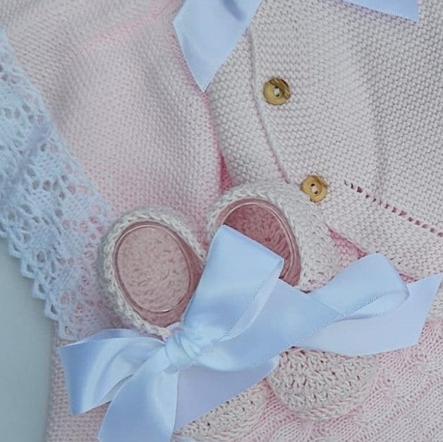A close-up of a light pink knitted baby outfit with white lace trim, gold buttons, and a large white satin bow, alongside matching white ribbon details.